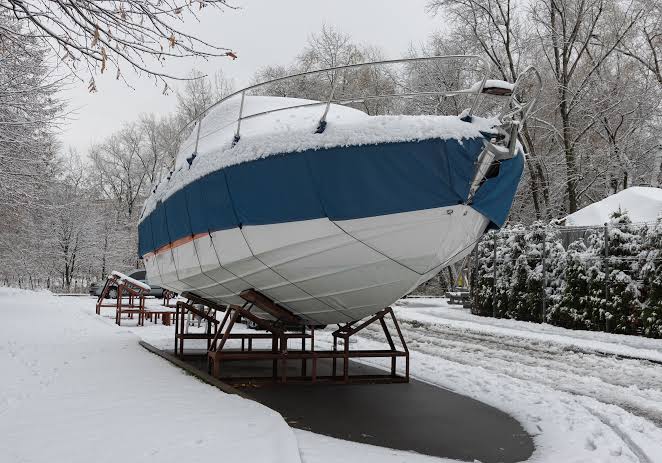 Snow-covered motorboat under a blue and white winter cover on storage stands outdoors